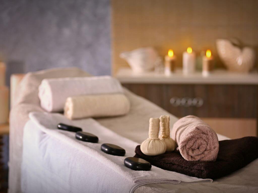 A picture of a spa treatment table with hot stones along one side, and rolled up towels at the head of the table. In the background are lit candles on a chest of drawers.