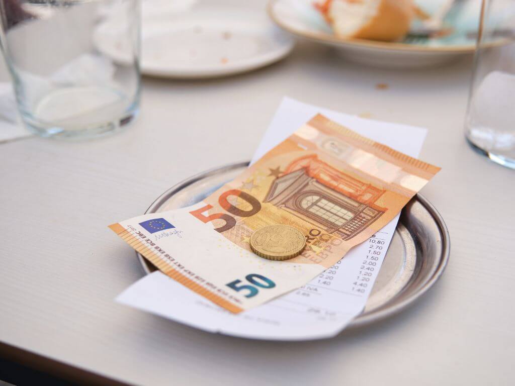 A picture of a small silver plate on a table that has a white table cloth. The silver plate has a food bill and on top of the bill is a fifty euro note and a fifty cent coin.