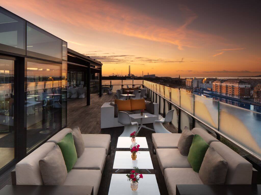 A picture of the Anantara Marker Hotel Rooftop Terrace in Dublin with tables and chairs n the terrace, the stacks of Ringsend visible in the background and the sky appears to be a sunset sky.