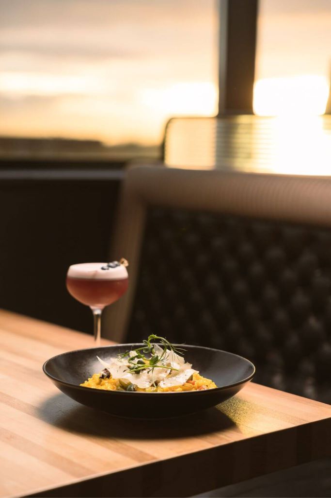 A picture of a meal and cocktail on a table at Sophie's Rooftop Bar at The Dean Hotel in Dublin with the sunset just visible in the background.
