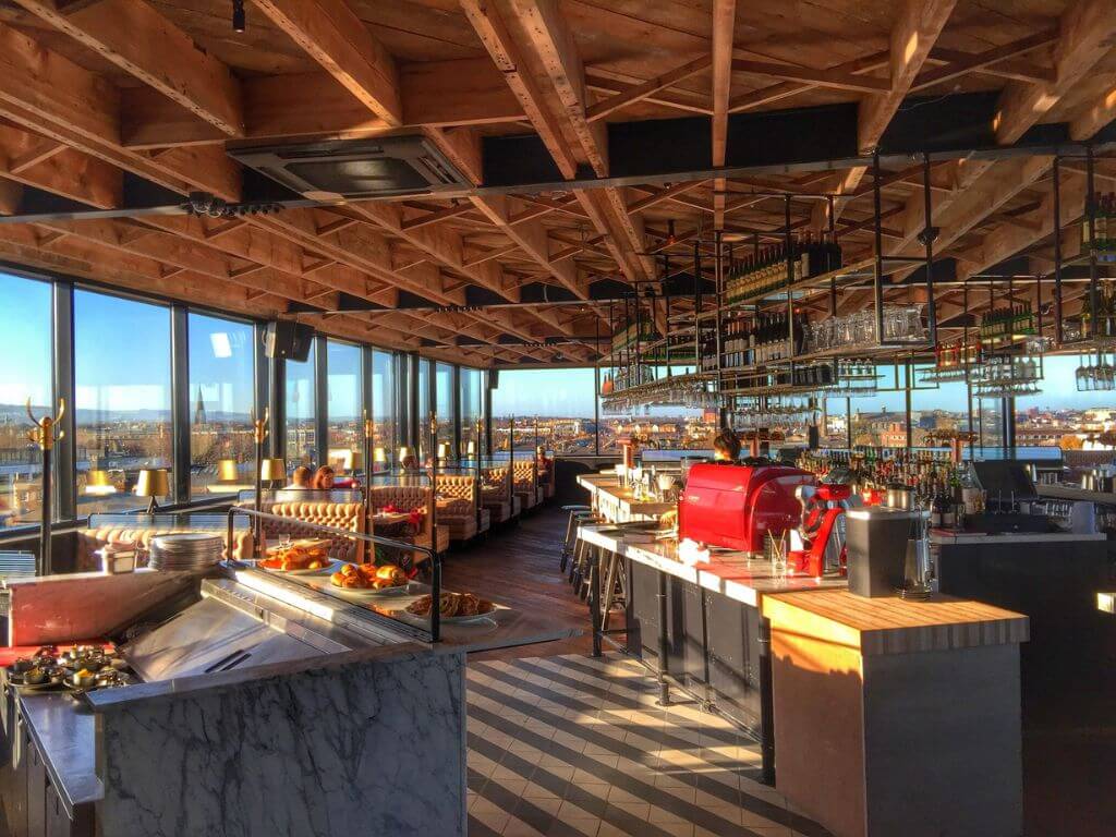 A picture of Sophie's Rooftop Bar at The Dean Hotel in Dublin with sun streaming in through the glass floor-to-ceiling windows and the wooden ceiling also visible.