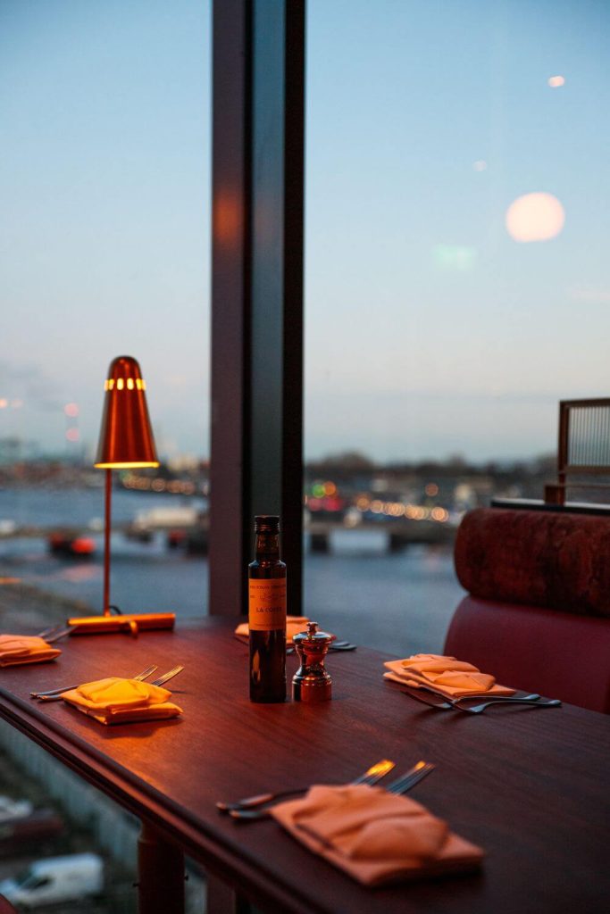 A picture of one of the tables at Ryleigh's at The Mayson Hotel, Dublin beside a window with a view over the river Liffey and a small table lamp illuminating the table.