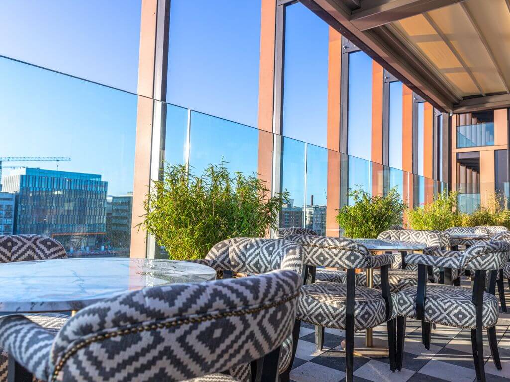 A picture of some of the round table with checkered white and black charis beside the terrace glass walls of Ryleigh's Rooftop Bar at The Mayson Hotel, Dublin with the city skyline visible in the background.