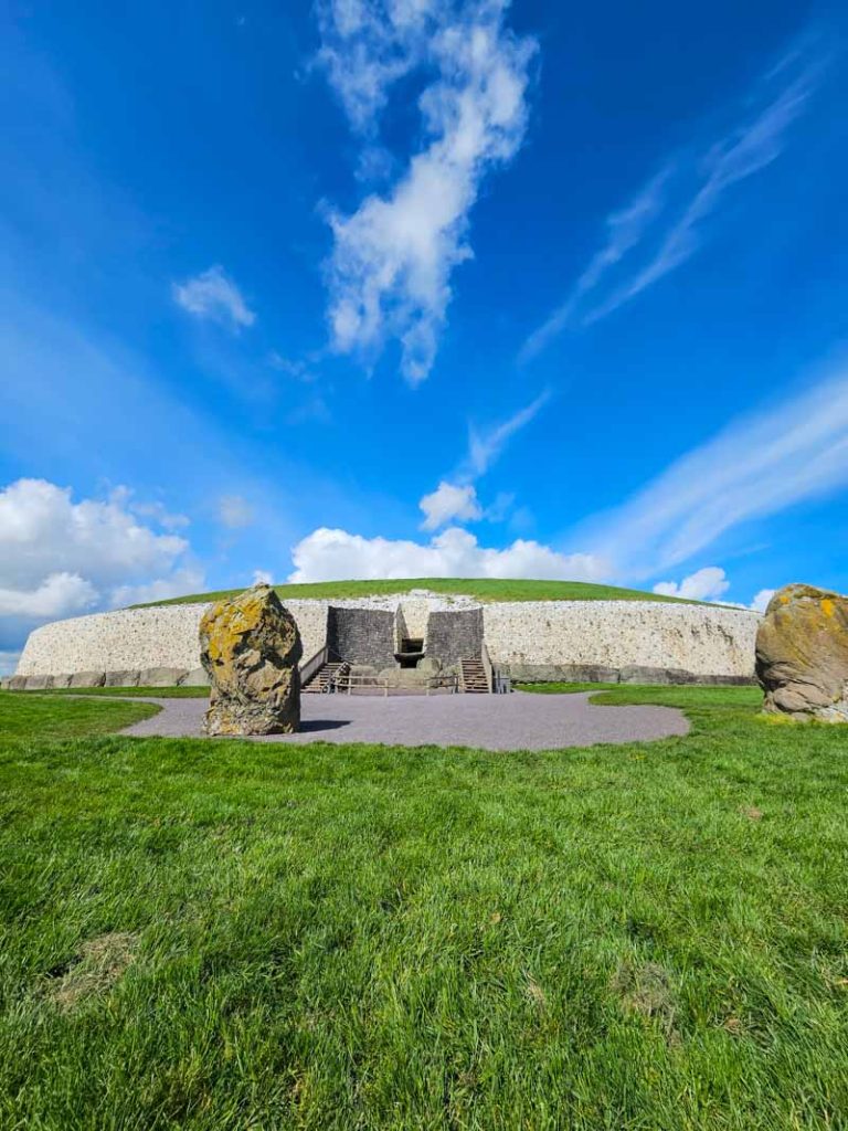 A picture of the mound of Newgrange Neolithic Passage Tomb with standing stones in front and blue skies with wispy clouds above.