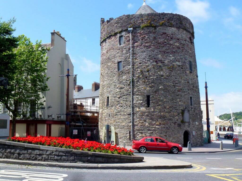 A picture of Reginald's Tower, Waterford City on a sunny day with a red car passing in front and red flowers in a banking in the foreground.