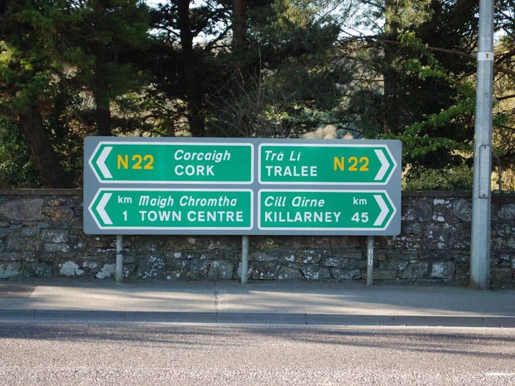 A picture of four road signs in Ireland, two pointing left and two pointing right, showing that they are in both English and Irish.