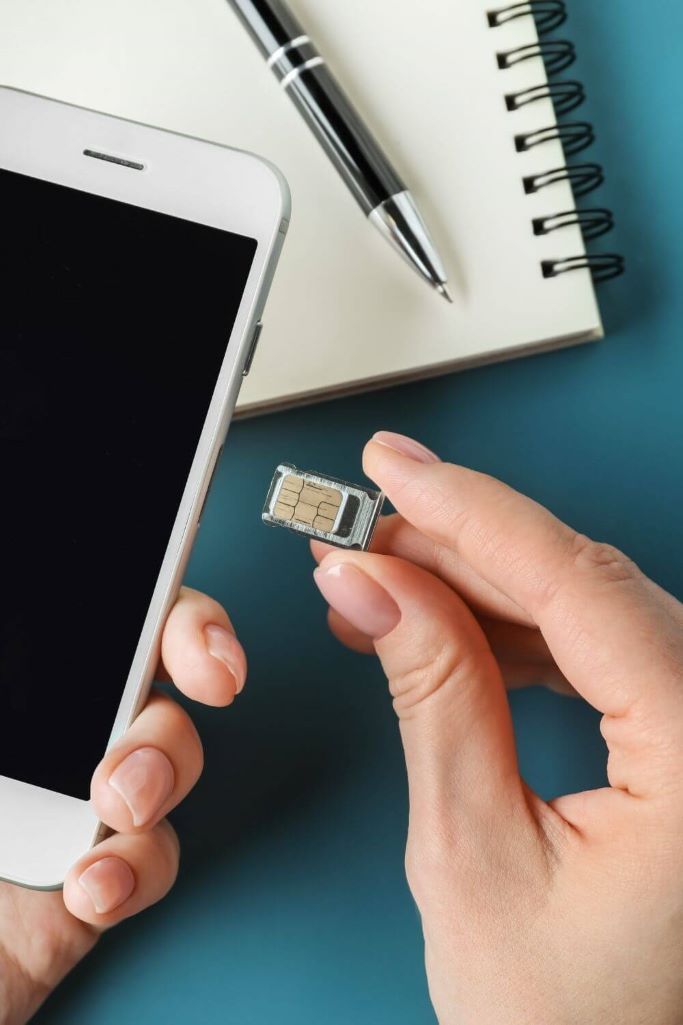 A picture of a person's hand holding a sim card towards an iPhone with a notebook and pen on the desk in the background.