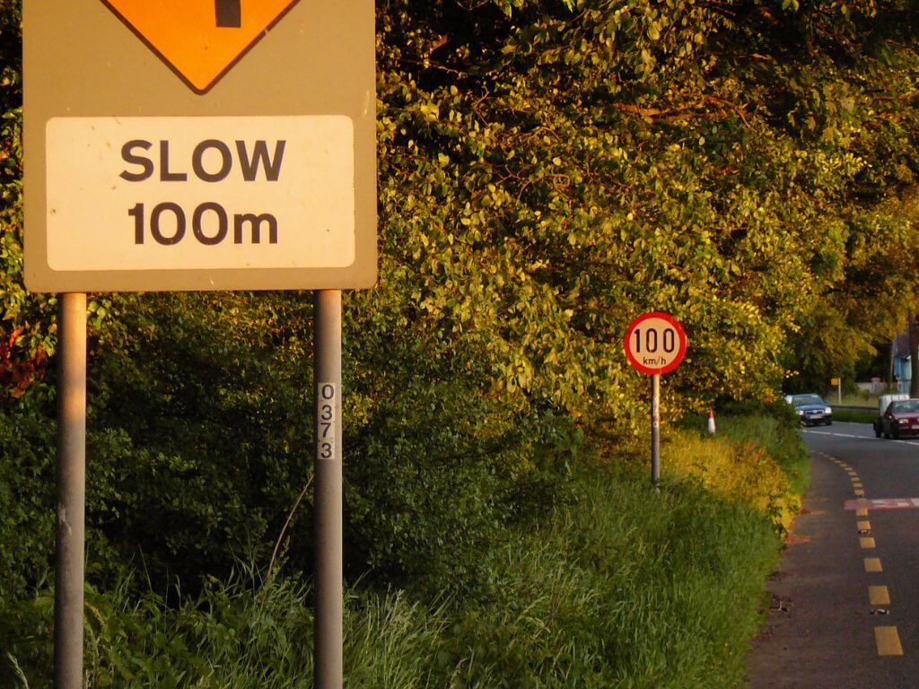 A picture of a 100km/h sign in Ireland along a road with a slow 100m sign before it.