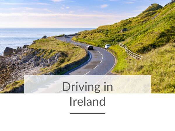 A picture of a winding road in Ireland running along the coast, with one car parked on the right of the road and one car passing on the left. There is text overlay at the bottom of the image saying driving in Ireland.