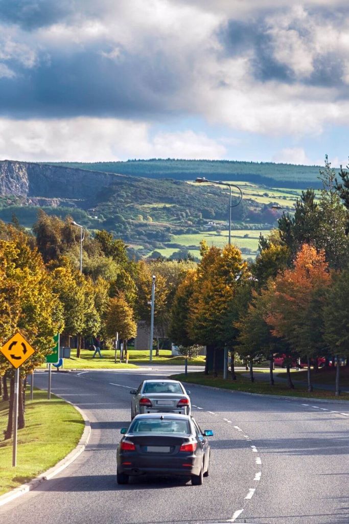 A picture of two cars driving on the left in Dublin, Ireland. The Dublin mountains are in the background and the road approaches a roundabout and is lined with trees.