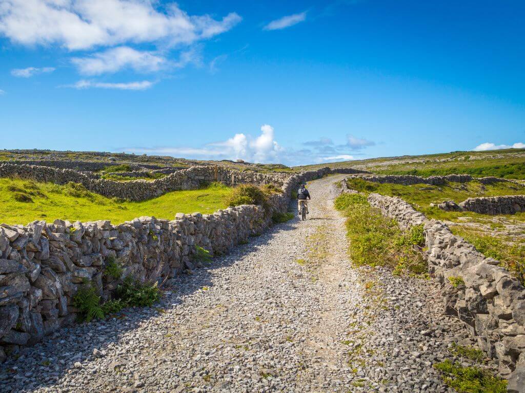 A picture of someone cycling along a gravel road on the Aran Islands in Ireland with stone walls on either side and blue skies overhead.