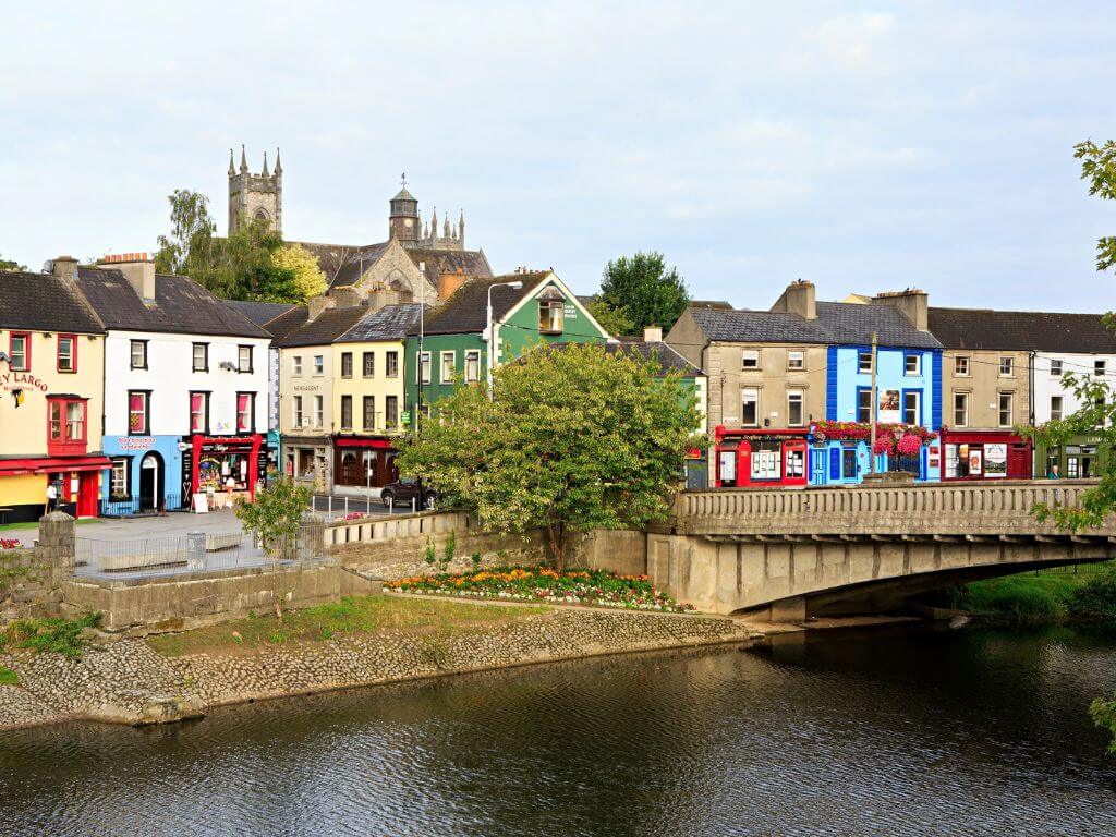 A picture of some of the colourful shop fronts along the river in Kilkenny, Ireland with the cathedral just visible over the building tops.