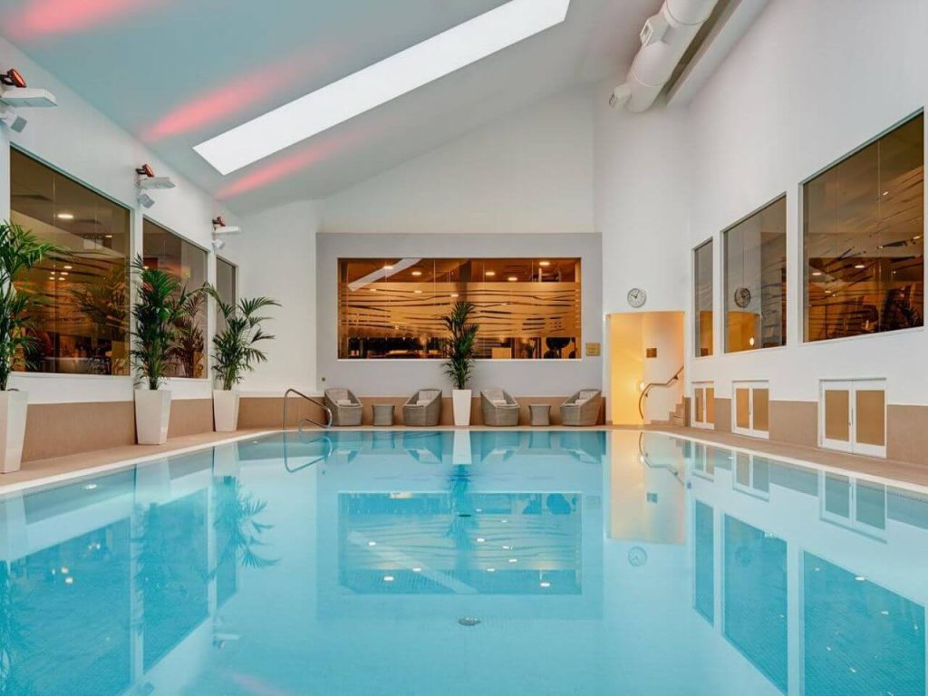 A picture of the indoor pool of The Shelbourne Hotel in Dublin Ireland. There are some rattan furniture in the background and frosted windows to either side of the pool.