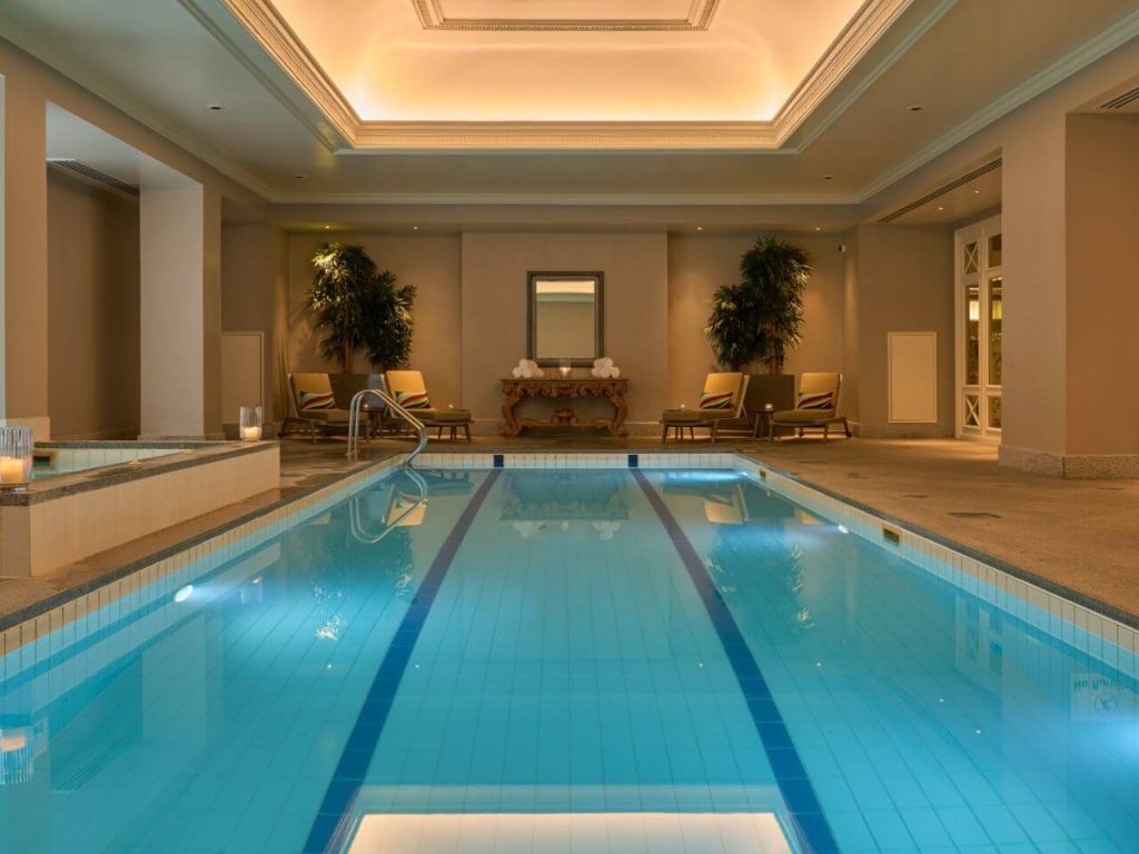 A picture of the indoor pool at the Intercontinental Four Seasons Hotel in Dublin, Ireland. There are four loungers and an ornate table with a mirror in the background and the edge of a jacuzzi to the left hand side of the pool.