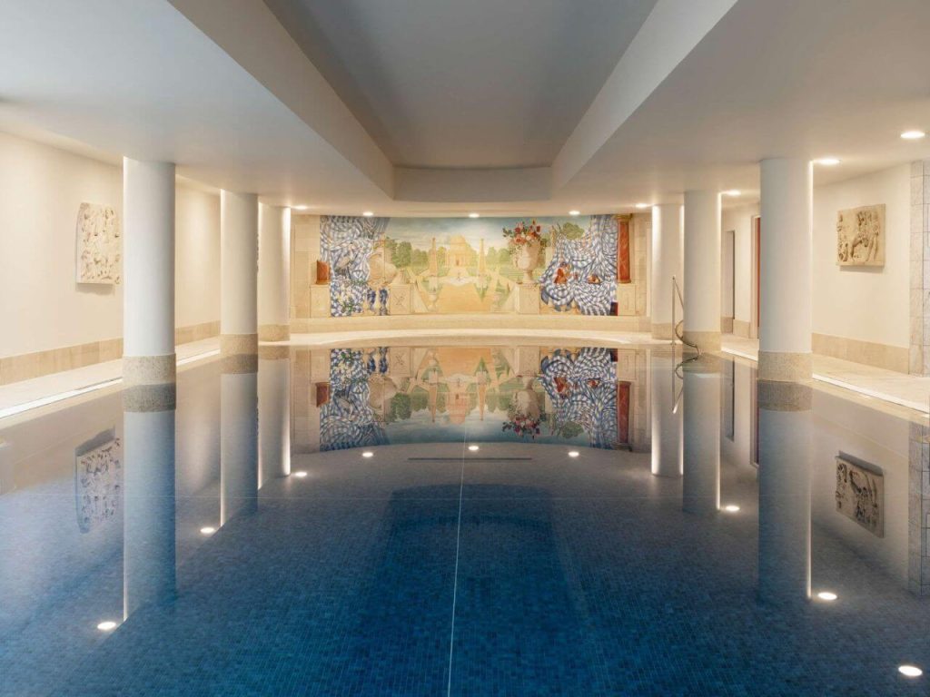 A picture of the indoor pool of the Merrion Hotel in Dublin. The pool surface is still, reflecting the columns on either side of the pool and the mural at the far end of the pool.
