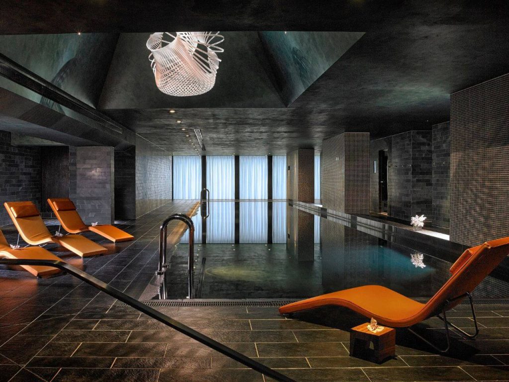 A picture of the indoor pool at The Anantara Marker Hotel in Dublin. It has orange reclining loungers around the pool, dark floor tiles and curtained windows in the background.