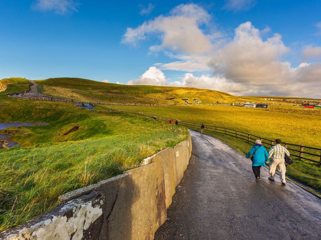 A picture of one of the pathways along the top of the Cliffs of Moher, with people wrapped up warm walking the path on a day with blue skies and a few white clouds overhead.