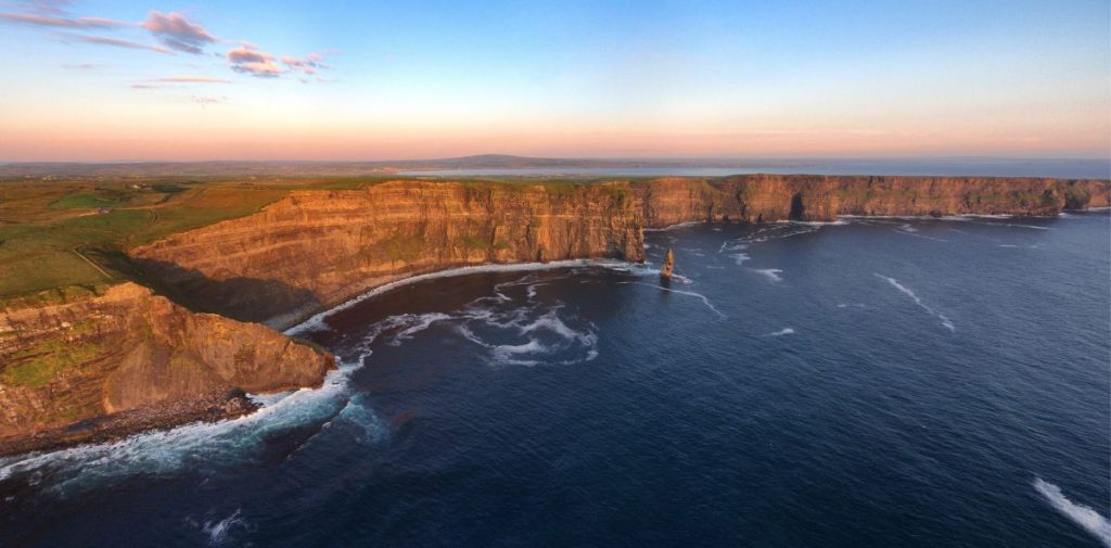 A stretched landscape picture of the Cliffs of Moher in County Clare in Ireland. There are waves breaking along the edge of the cliffs and there are some hills just visible in the far background. The cliffs are stretching from left to right.